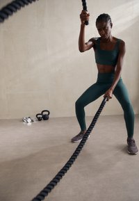 Un individu en tenue de sport vert foncé saisit une corde de combat noire. Un ensemble de poids est visible en arrière-plan sur une surface claire.