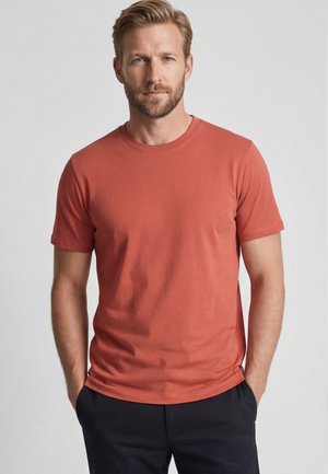 Blond man met baard, met een effen roestkleurig T-shirt met korte mouwen en donkere broek, staand met handen in de zakken tegen een grijze achtergrond.