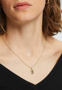 Goldkette mit rechteckigem Anhänger, der eingravierte Sternenmuster zeigt, befestigt an einer feinen Kette, getragen über einem schwarzen V-Ausschnitt-Oberteil.