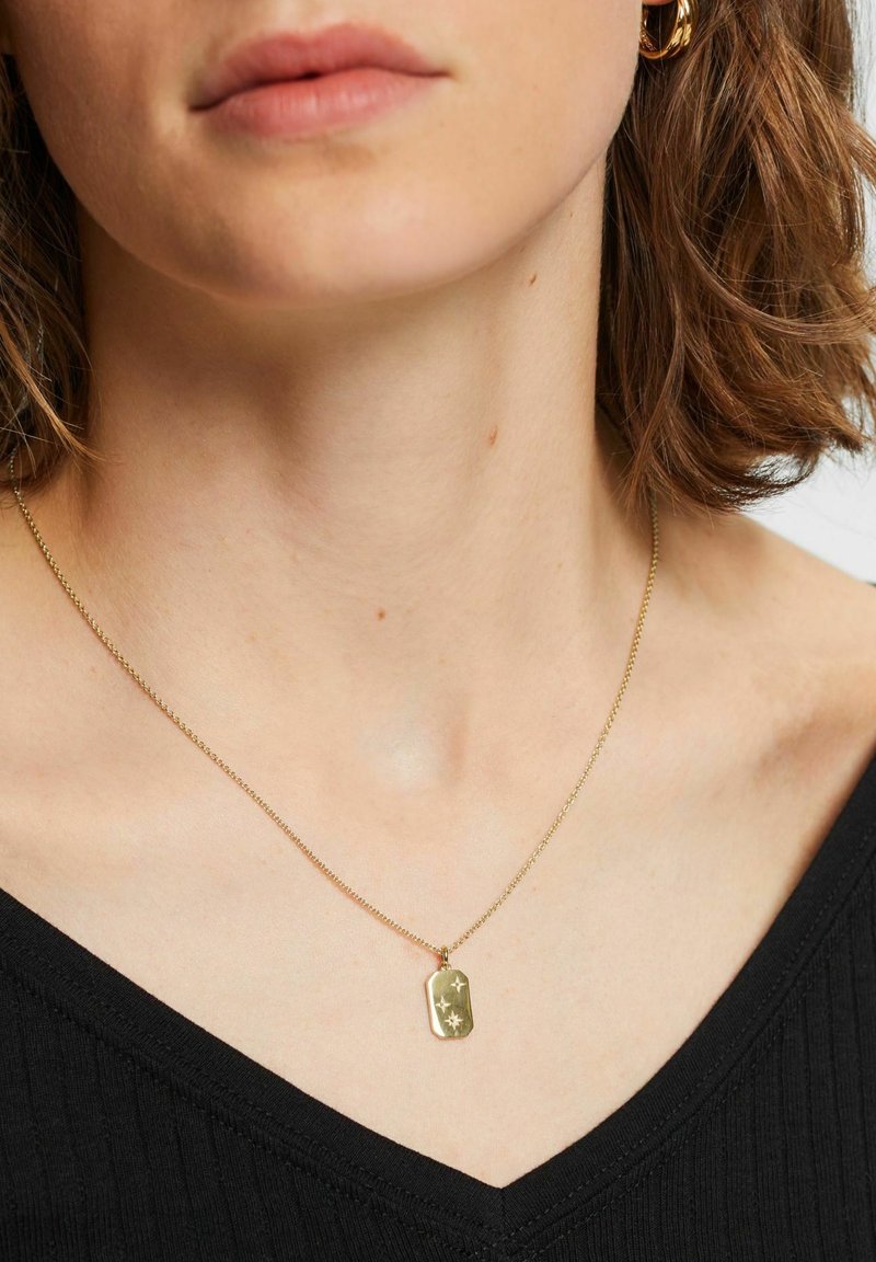 Goldkette mit rechteckigem Anhänger, der eingravierte Sternenmuster zeigt, befestigt an einer feinen Kette, getragen über einem schwarzen V-Ausschnitt-Oberteil.