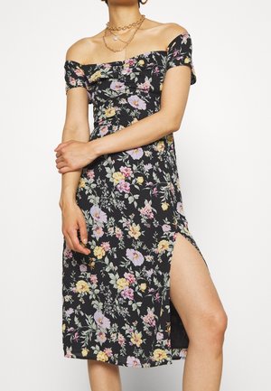 Mujer con vestido negro floral con hombros descubiertos, mangas cortas, abertura lateral y collares de cadena dorada en capas sobre fondo blanco.