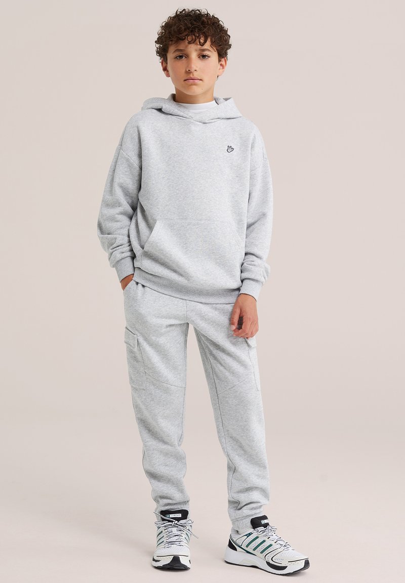 Garçon aux cheveux bouclés, portant un sweat à capuche gris clair, un pantalon de survêtement assorti et des chaussures de sport blanches avec des détails verts et noirs.
