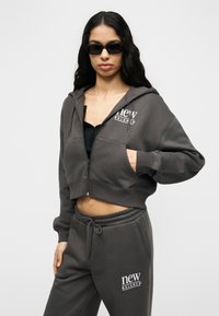 Γυναίκα που φοράει σκουρόγρινη κοντή μπλούζα με κουκούλα και αντίστοιχο παντελόνι φόρμας New Balance, με γυαλιά ηλίου και το χέρι στην τσέπη, στέκεται μπροστά σε λευκό φόντο.