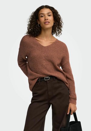 Femme aux cheveux bouclés portant un pull en tricot marron et un pantalon foncé, tenant un sac à main noir d'une main sur un fond uni.