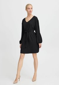 Robe noire texturée avec un décolleté en V profond, des manches bouffantes longues et une taille cintrée par un nœud. Associée à des talons hauts beiges.