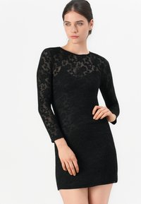 Robe en dentelle noire avec des manches longues, un motif floral, une silhouette cintrée et un col rond. La texture semble transparente avec une finition lisse.