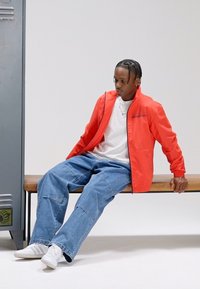 Röd lättviktsjacka, vit t-shirt, blå baggy jeans och vita sneakers. Sitter på en träbänk bredvid ett metallskåp.