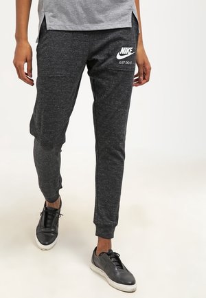 Personne portant un jogging Nike gris foncé avec logo et slogan « Just Do It », associé à des baskets noires et un t-shirt gris clair.
