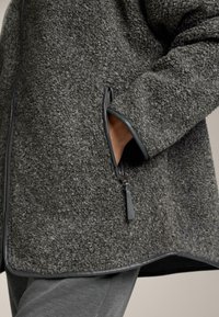 Siva flis jakna z teksturirano površino, z zadrgami na žepih in črnimi obrobami vzdolž robov, prikazana delno odprta.