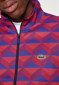Veste zippée multicolore avec un motif géométrique de cubes rouges et bleus, avec un petit logo vert Lacoste sur la poitrine.