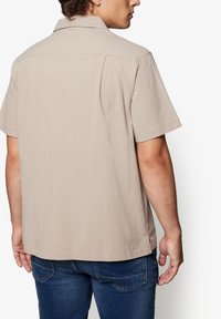Short-sleeve beige shirt with a textured fabric, button-down collar, and back shoulder seams. Paired with blue denim jeans.