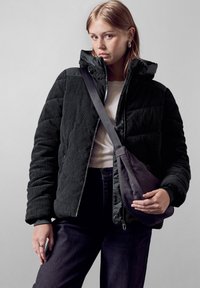 Giacca puffer in corduroy nera con cappuccio, chiusura con zip frontale, tasche laterali e una fodera nera liscia, abbinata a una borsa a tracolla grigia.