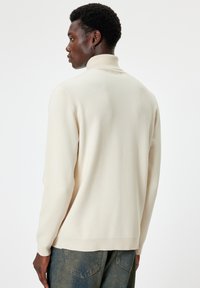 Kremowy sweter z golfem i prążkowanymi detalami, wykonany z miękkiej dzianiny. Cechuje się luźnym krojem i prostym designem, włożony w niebieskie dżinsy.