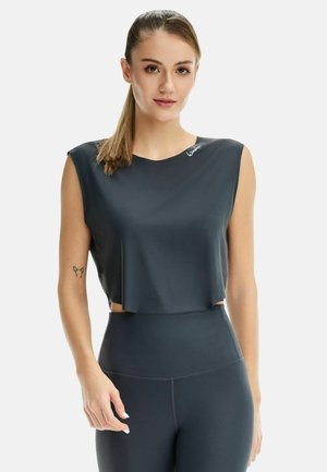 Young woman wearing a sleeveless cropped black athletic top and high-waisted leggings with hair in a ponytail against white background.