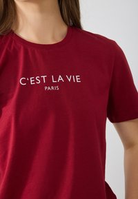 T-shirt en coton rouge avec un col rond et des manches courtes. Le texte "C'EST LA VIE PARIS" est imprimé en blanc. Texture douce, design décontracté.