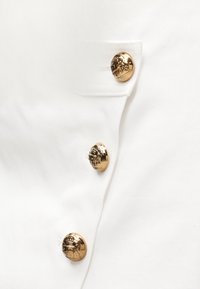 Λευκό ύφασμα με μια σειρά από τρία λαμπερά χρυσά κουμπιά και μια μικρή λεπτομέρεια τσέ πης, που αναδεικνύει έναν ομαλό, δομημένο σχεδιασμό.