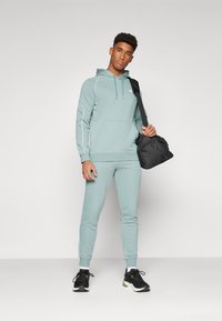 Haut à capuche vert clair avec une poche avant et un passepoil blanc, associé à un jogging assorti. Chaussures de sport noires et sac noir.