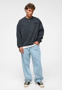 Donkergrijze oversized sweatshirt met een textuurafwerking, gedragen over een wit shirt, gecombineerd met lichtblauwe baggy jeans en zwarte sneakers.