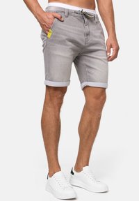 Shorts en denim gris avec des revers, poches avant et une étiquette jaune contrastante à la taille. Accompagnés de baskets blanches.