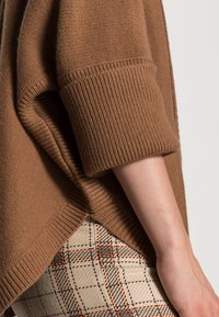 Bras portant un pull marron tricoté retroussé et un pantalon à carreaux avec des lignes beige, noir et orange sur un fond clair.
