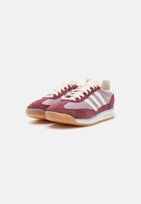 Mahognyfärgade mockaskor med krämfärgade snören och detaljer. Har tre vita ränder och en blå detalj på sulan. Texturerad gummisula.