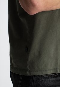 Zijaanzicht van een persoon in een olijfgroene T-shirt en donkere jeans, met de arm gebogen en over het lichaam gekruist.