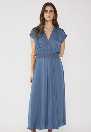 Donna in piedi che indossa un abito lungo blu con maniche corte, scollo a V, vita arricciata e gonna plissettata, su uno sfondo chiaro uniforme.