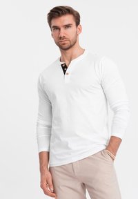 Valkoinen pitkähihainen Henley-paita, jossa on musta napitus, kolme nappia ja pehmeä kangas. Yhdistetty vaaleanbeigeihin housuihin.