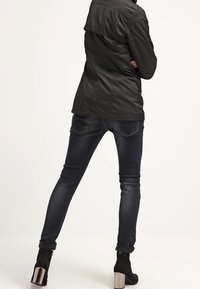 Svart, lättviktsjacka med krage och flik, kombinerad med mörka jeans och ankelstövlar med metalliska detaljer, sedd bakifrån.