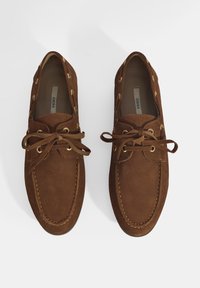 Braune Wildlederschuhe mit runder Zehenpartie, Schnürdesign, goldenen Ösen und gestickten Akzenten entlang der Sohle und des Schafts.
