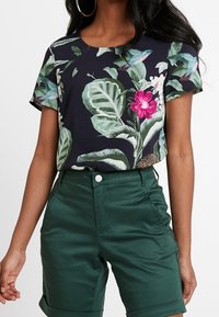 Koszulka z kwiatowym nadrukiem w kolorze granatowym, z zielonymi liśćmi i różowymi kwiatami, zestawiona z zielonymi szortami o wysokim stanie, z zapięciem na guzik i czterema kieszeniami.