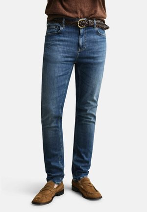 Person trägt eng anliegende blaue Jeans, braunen geflochtenen Gürtel, braunes Langarmhemd und braune Wildleder-Loafer und steht vor weißem Hintergrund.