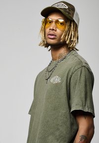 T-shirt surdimensionnée vert olive avec un logo graphique, collier en chaîne argentée superposé, casquette à motif camouflage et lunettes de soleil aviateur teintées jaunes.