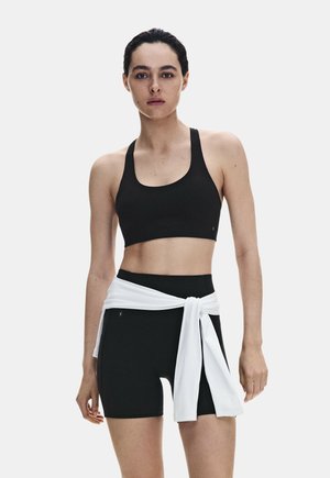 Frau trägt einen schwarzen Sport-BH und schwarze Radlerhosen mit einem weißen Langarmshirt, das um die Taille gebunden ist, und steht vor einem schlichten Hintergrund.