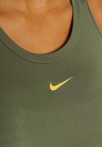 Detailní záběr osoby v olivově zeleném tílku s žlutým logem Nike swoosh na hrudi.