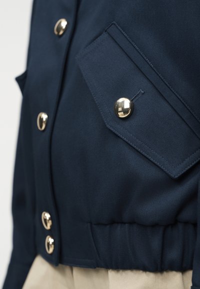 Κοντινό πλάνο της τσέπης ενός σκούρου μπλε σακακιού με χρυστό κουμπί στο καπάκι, επιπλέον χρυστά κουμπιά μπροστά και ελαστική ζώνη στη μέση.