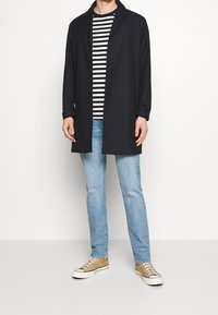 Laivan sininen pitkä takki päällä raidoitetun paidan, vaaleansinisten farkkujen ja beigeltä näyttävien tennareiden kanssa. Takissa on lohkottu kaulus ja kaksi nappia.