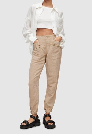 Kobieta ubrana w beżowe spodnie z wysokim stanem i zapinane kieszenie, biały krótki top, otwartą białą koszulę oraz czarne masywne sandały.