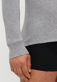 Top gri cu mânecă lungă, cu textură cu ribbing, având un design ajustat și mâneci cu manșete, purtat cu pantaloni scurți negri.