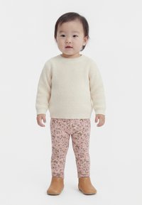 Kleinkind steht, trägt einen cremefarbenen Strickpullover, pinke Leggings mit Blumenmuster und hellbraune Stiefeletten, vor einem schlichten weißen Hintergrund.