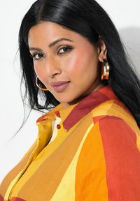 Una camicia a righe colorate che presenta tonalità di giallo, arancione e rosso; include un colletto e dettagli con bottoni. Orecchini a cerchio grandi completano il look.