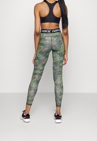 Donna vista di spalle che indossa un reggiseno sportivo nero, leggings verdi con fantasia e una vita con logo Nike, e sneakers Nike bianche e nere.