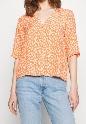 Femme portant un chemisier orange à manches courtes avec un motif floral blanc et un jean taille haute bleu clair, debout devant un fond uni.