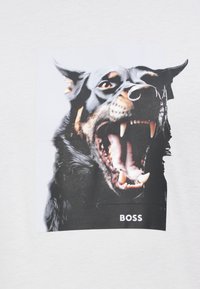 Λευκό μπλουζάκι με μεγάλο σχέδιο που απεικονίζει το έντονο πρόσωπο ενός μαύρου σκύλου, με φανερά δόντια και δαχτυλίδι στη μύτη. Το κείμενο από κάτω γράφει "BOSS."