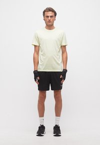 Νεαρός άνδρας που στέκεται, φοράει ανοιχτό κίτρινο μπλουζάκι, μαύρο σορτς, μαύρα γάντια χωρίς δάχτυλα, λευκές κάλτσες και μαύρα παπούτσια τρεξίματος μπροστά σε λευκό φόντο.