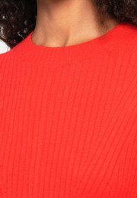 Gros plan d'une personne portant un pull en maille côtelée rouge vif avec un col rond.