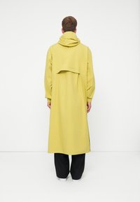 Impermeável amarelo longo com capuz, apresentando um design em camadas nas costas e mangas longas. Combinado com calças pretas e sapatos brancos.