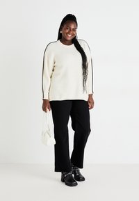 Λευκό πλεκτό πουλόβερ με μαύρες λεπτομέρειες στα πλάγια, μαύρο παντελόνι με φαρδιά πόδια, μαύρες chunky παπούτσια με τακούνι, και μια μικρή λευκή τσάντα.