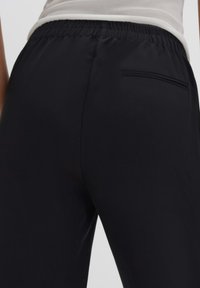 Zwarte elastische taillebandbroeken met een gladde textuur, voorzien van een achterzak met een rechte lijnaccent. Eenvoudig en ingetogen ontwerp.