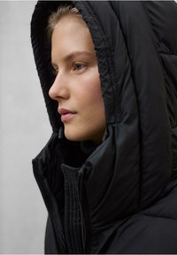 Mladá žena se světlou pletí a modrýma očima má na sobě černou bundu s kapucí a stojí nalevo na jednobarevném šedém pozadí.